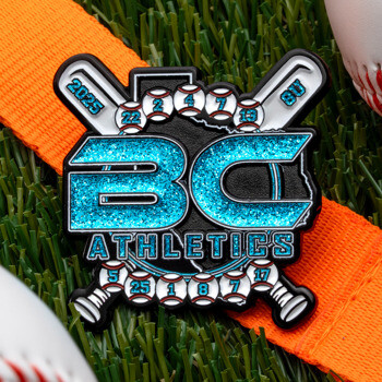 BC Athletics baseball lapel pin with glitter teal lettering and crossed bats, resting on an orange strap in grass near baseballs.