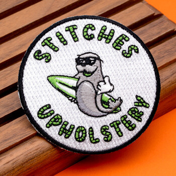 Circular embroidered patch for Stitches Upholstery featuring a sea otter wearing sunglasses and holding a surfboard.