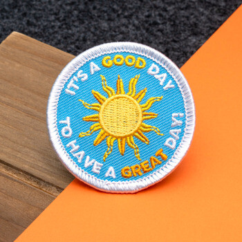 Circular embroidered patch featuring a yellow sun on a light blue background with white and yellow text.