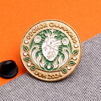 Round gold enamel lapel pin with green and white details featuring an anatomical heart and text Donor Champion UUH 2024.