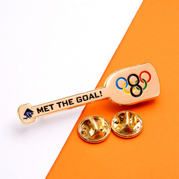 Gold-toned oar-shaped lapel pin with Olympic rings and Met The Goal text, shown with two butterfly clutch backings.