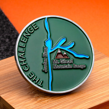 Round green challenge coin featuring the Jordan River, Mt. Gilead Mountain Range, and The Challenge text resting on a wooden block.