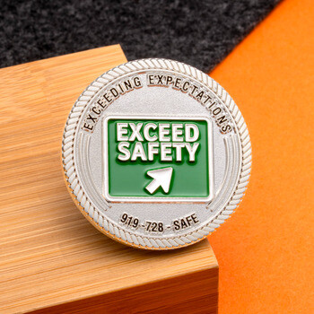 Silver challenge coin featuring a green Exceed Safety sign logo with rope cut edge resting on a wooden block.