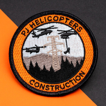 Circular embroidered patch for PJ Helicopters Construction featuring utility helicopters, a power transmission tower, mountains, and an orange border.